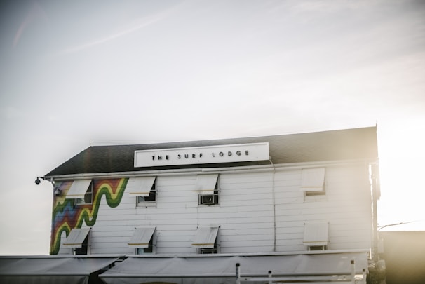 A white building with a sign that reads 'The Surf Lodge' is shown. The exterior features multiple windows with white shutters. A colorful wave design is seen painted on one side of the building. The sky is clear with gentle sunlight creating a serene atmosphere.