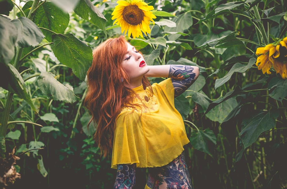 a woman with red hair and tattoos standing in a field of sunflowers