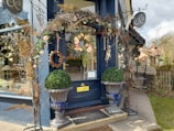 The entrance of a quaint boutique is adorned with a floral archway made of dried branches and colorful flowers, including roses and leaves. Two symmetrical large planters with spherical topiary rest in front of a deep blue-painted storefront. A sign for the shop hangs above, and through the window, decorative items are visible, suggesting a focus on interior decor.