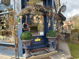 The entrance of a quaint boutique is adorned with a floral archway made of dried branches and colorful flowers, including roses and leaves. Two symmetrical large planters with spherical topiary rest in front of a deep blue-painted storefront. A sign for the shop hangs above, and through the window, decorative items are visible, suggesting a focus on interior decor.