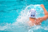 A water polo player wearing a white cap, identified by the number 6, is in the middle of a dynamic action, splashing water around while swimming in a pool. The player is oriented with their back visible, showcasing muscular shoulders.