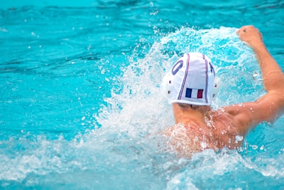 A water polo player wearing a white cap, identified by the number 6, is in the middle of a dynamic action, splashing water around while swimming in a pool. The player is oriented with their back visible, showcasing muscular shoulders.