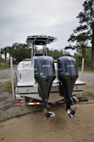 A boat is parked on a trailer with two powerful Yamaha outboard engines at the rear. The setting appears to be an outdoor area with trees and a cloudy sky in the background.