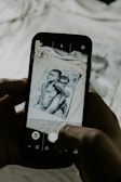 A smartphone is held in two hands capturing a photo of an image. The image on the phone screen, as well as partially visible in the background, depicts two children. The photograph aligns with the phone's camera interface features, including buttons and icons on the screen.