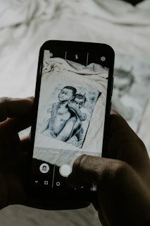 A smartphone is held in two hands capturing a photo of an image. The image on the phone screen, as well as partially visible in the background, depicts two children. The photograph aligns with the phone's camera interface features, including buttons and icons on the screen.