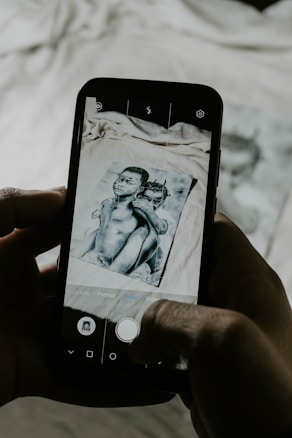 A smartphone is held in two hands capturing a photo of an image. The image on the phone screen, as well as partially visible in the background, depicts two children. The photograph aligns with the phone's camera interface features, including buttons and icons on the screen.