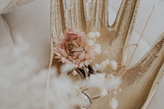 A gold decorative hand sculpture holds a pink dried flower and several silver rings. Delicate white dried flowers are scattered around, creating a soft, elegant composition.