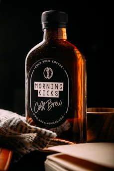 A bottle labeled 'Morning Kicks Cold Brew' is placed on a surface with dim lighting. The bottle has a dark label with text and is filled with a dark liquid, likely coffee. There is a piece of patterned fabric draped near the bottom of the bottle and a wooden object next to it.