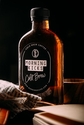 A bottle labeled 'Morning Kicks Cold Brew' is placed on a surface with dim lighting. The bottle has a dark label with text and is filled with a dark liquid, likely coffee. There is a piece of patterned fabric draped near the bottom of the bottle and a wooden object next to it.