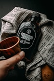 A bottle labeled 'Morning Kicks Cold Brew' placed on a textured, printed cloth. A hand is holding a small wooden cup near the bottle, creating a rustic and cozy atmosphere.
