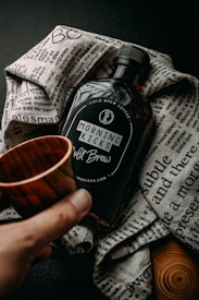 A bottle labeled 'Morning Kicks Cold Brew' placed on a textured, printed cloth. A hand is holding a small wooden cup near the bottle, creating a rustic and cozy atmosphere.