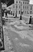 A street scene features a few people navigating wet and icy conditions. Lampposts line the sidewalk, and footprints are visible on the slushy ground. Umbrellas are used to shield from ongoing precipitation, which appears to be a mix of rain and hail.