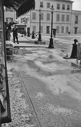A street scene features a few people navigating wet and icy conditions. Lampposts line the sidewalk, and footprints are visible on the slushy ground. Umbrellas are used to shield from ongoing precipitation, which appears to be a mix of rain and hail.
