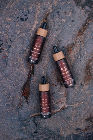 Three dark brown bottles with wooden caps and dropper tops are placed on a rough, textured gray surface. The bottles have labels with text indicating different product names.