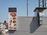 A signboard displaying a variety of fast food items such as burgers, wraps, fries, pretzels, and coffee. The sign indicates the establishment is open and is attached to a building with metal siding. The sign contains the text 'K.I. Imbiss & Bäckerei'. The background shows a clear blue sky and part of a neighboring building.