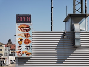 A signboard displaying a variety of fast food items such as burgers, wraps, fries, pretzels, and coffee. The sign indicates the establishment is open and is attached to a building with metal siding. The sign contains the text 'K.I. Imbiss & Bäckerei'. The background shows a clear blue sky and part of a neighboring building.