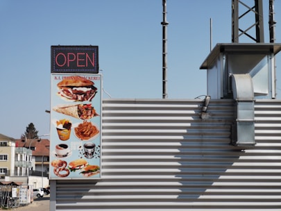 A signboard displaying a variety of fast food items such as burgers, wraps, fries, pretzels, and coffee. The sign indicates the establishment is open and is attached to a building with metal siding. The sign contains the text 'K.I. Imbiss & Bäckerei'. The background shows a clear blue sky and part of a neighboring building.