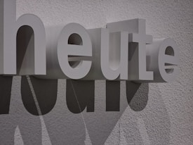 Large white 3D letters spell out the word 'heute' against a textured wall. The letters cast distinct shadows, adding depth and interest to the composition.