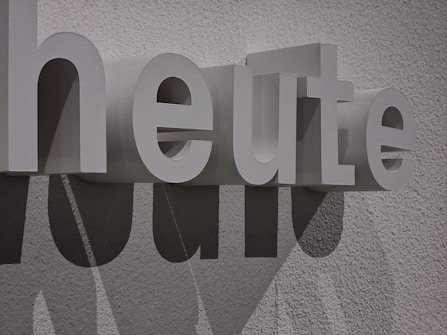 Large white 3D letters spell out the word 'heute' against a textured wall. The letters cast distinct shadows, adding depth and interest to the composition.