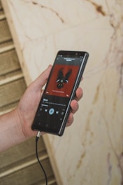 A hand holding a smartphone with an album cover displayed on the screen. The album cover features a stylized image of a bird on a red background. The phone is connected to headphones via a jack.
