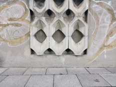 A textured concrete wall with decorative geometric cutouts is adorned with graffiti. The wall features symmetrical, diamond-shaped designs repeated across its surface. The ground is paved with large rectangular tiles, one of which has an object that looks like a small piece of debris or litter.