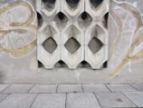 A textured concrete wall with decorative geometric cutouts is adorned with graffiti. The wall features symmetrical, diamond-shaped designs repeated across its surface. The ground is paved with large rectangular tiles, one of which has an object that looks like a small piece of debris or litter.