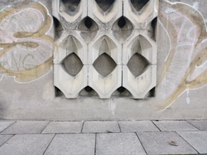 A textured concrete wall with decorative geometric cutouts is adorned with graffiti. The wall features symmetrical, diamond-shaped designs repeated across its surface. The ground is paved with large rectangular tiles, one of which has an object that looks like a small piece of debris or litter.