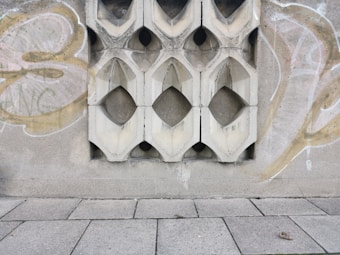 A textured concrete wall with decorative geometric cutouts is adorned with graffiti. The wall features symmetrical, diamond-shaped designs repeated across its surface. The ground is paved with large rectangular tiles, one of which has an object that looks like a small piece of debris or litter.