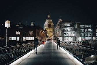 A nighttime scene featuring a pedestrian bridge leading towards a large, illuminated domed building in the background. Several people are walking along the bridge, with the city buildings flanking the sides. The surrounding structures, including the City of London School, are visible and appear to be partially lit.
