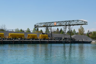 An industrial setting by the water featuring a series of yellow machinery or containers aligned on a structure, possibly used for loading or transportation. There's a large pile of gravel or similar material, with a metal framework overhead suggesting a conveyor system. Trees and foliage can be seen in the background under a clear blue sky.