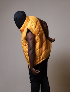 A person with a black covered face is wearing a sleeveless puffy orange vest and dark pants. The individual is bending slightly forward against a neutral background, creating a concealed and mysterious appearance.