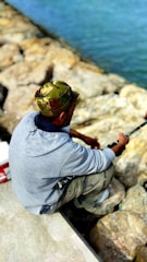 A person wearing a camouflage cap and a gray sweatshirt is sitting on a rocky shoreline, holding a fishing rod. The rocks are large and light-colored, while the water appears blue and calm.