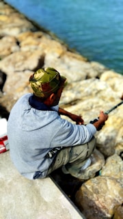 A person wearing a camouflage cap and a gray sweatshirt is sitting on a rocky shoreline, holding a fishing rod. The rocks are large and light-colored, while the water appears blue and calm.
