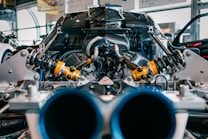 A close-up view of an intricate car engine, focusing on the suspension and shock absorber parts. The image captures the metallic and mechanical components, highlighting the yellow and black shock assemblies against a backdrop of carbon fiber and other engine elements.