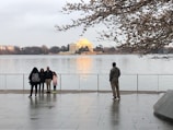 A group of people stands by a waterfront with the Jefferson Memorial in the background. The people are facing the water, some are posing for a picture while one person stands separately. The scene is serene with cherry blossom branches partially framing the top of the image.