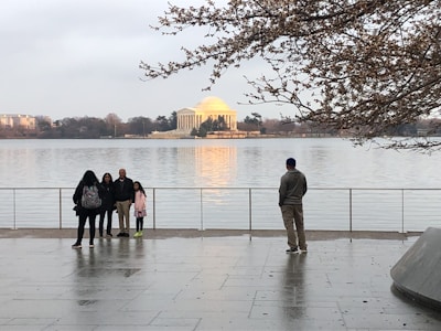 A group of people stands by a waterfront with the Jefferson Memorial in the background. The people are facing the water, some are posing for a picture while one person stands separately. The scene is serene with cherry blossom branches partially framing the top of the image.