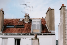 A rooftop characterized by red clay tiles and several chimneys. There are two skylights and an old-style television antenna perched on the roof. The wall beneath the roof is white, and the sky above is partly cloudy.