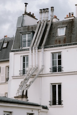 A rooftop scene featuring a series of tall, narrow chimneys extending from a modern building with a slate roof. The structure is composed of multiple sections, each topped with its own chimney stack. The sky above is overcast, casting a pale light on the architecture.