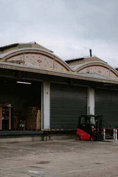 A loading dock area with closed roller doors, numbered 5 and 6. There are stacks of cardboard boxes visible in an open section of the dock, and a red and black forklift is parked nearby. The building features a facade of brick arch designs under a cloudy sky.