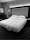 A neatly made bed with multiple pillows is positioned in a hotel room. The headboard is upholstered and complemented by wall lighting above it. A bedside table holds a phone, and a modern lamp stands next to an armchair. Decorative framed art adorns the wall, and a geometric patterned carpet covers the floor.