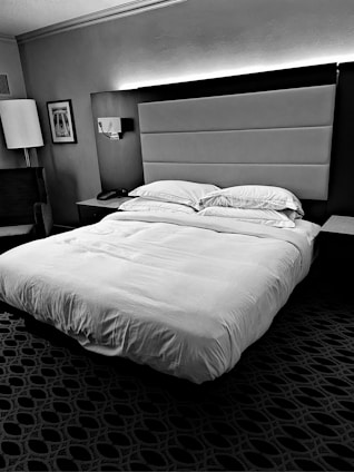 A neatly made bed with multiple pillows is positioned in a hotel room. The headboard is upholstered and complemented by wall lighting above it. A bedside table holds a phone, and a modern lamp stands next to an armchair. Decorative framed art adorns the wall, and a geometric patterned carpet covers the floor.