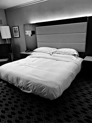 A neatly made bed with multiple pillows is positioned in a hotel room. The headboard is upholstered and complemented by wall lighting above it. A bedside table holds a phone, and a modern lamp stands next to an armchair. Decorative framed art adorns the wall, and a geometric patterned carpet covers the floor.