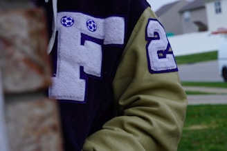 A close-up of a letterman jacket featuring a large letter 'F' with soccer ball designs and the number '24' on its sleeve. The jacket is two-toned, with dark and light fabric. A background of a residential area with grass is partially visible.