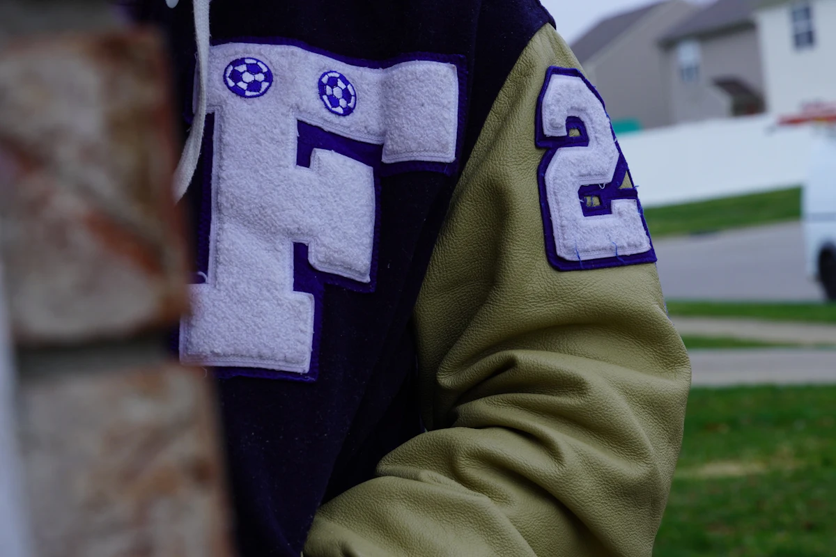 Detail patch chenille angka 62 dan huruf P warna krem di dada jaket varsity hitam, difoto dari jarak dekat