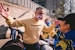 A person wearing a yellow sweatshirt with a phrase in Cyrillic is speaking to another person. The person has a mask with the British flag design. There are several others in the background, some wrapped in Ukrainian flags. The setting appears to be outdoors on a sunny day, with a concrete wall and a building visible.