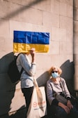 A person holds a flag with blue and yellow stripes in front of their face while standing against a light-colored brick wall. Another person, wearing sunglasses and a face mask, sits on a ledge below, holding a handbag. The seated person wears casual clothing and the standing individual carries a tote bag.
