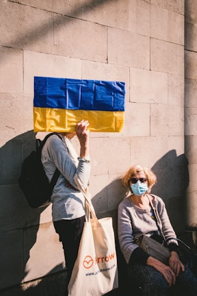 A person holds a flag with blue and yellow stripes in front of their face while standing against a light-colored brick wall. Another person, wearing sunglasses and a face mask, sits on a ledge below, holding a handbag. The seated person wears casual clothing and the standing individual carries a tote bag.