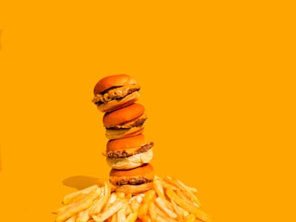 A stack of four cheeseburgers is balanced atop each other, surrounded by a scattered pile of golden French fries. The background is a solid bright yellow, enhancing the vibrant colors of the fast food items.