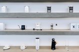 Shelves display various coffee brewing equipment and accessories. The top shelf holds tins labeled with coffee blends. The middle shelf showcases a black kettle, a coffee dripper with a branded cup, grinder, and a few boxes. The bottom shelf features a pair of coffee cups, a tall white thermos, a black object resembling a kettle, and an empty glass.