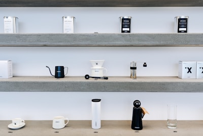 Shelves display various coffee brewing equipment and accessories. The top shelf holds tins labeled with coffee blends. The middle shelf showcases a black kettle, a coffee dripper with a branded cup, grinder, and a few boxes. The bottom shelf features a pair of coffee cups, a tall white thermos, a black object resembling a kettle, and an empty glass.